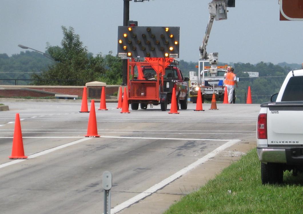 Roadside lane closure with orange cones, maintenance trucks, and a lifted worker crane on a highway shoulder