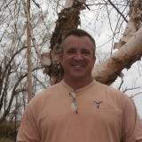 Smiling person in a tan shirt standing outdoors in front of leafless trees.