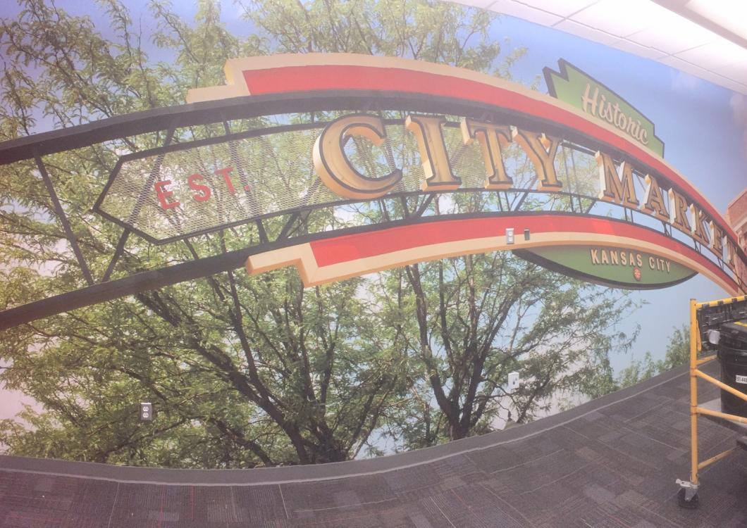 Curved colorful metal arch sign outdoors among trees, with a small ladder at right.