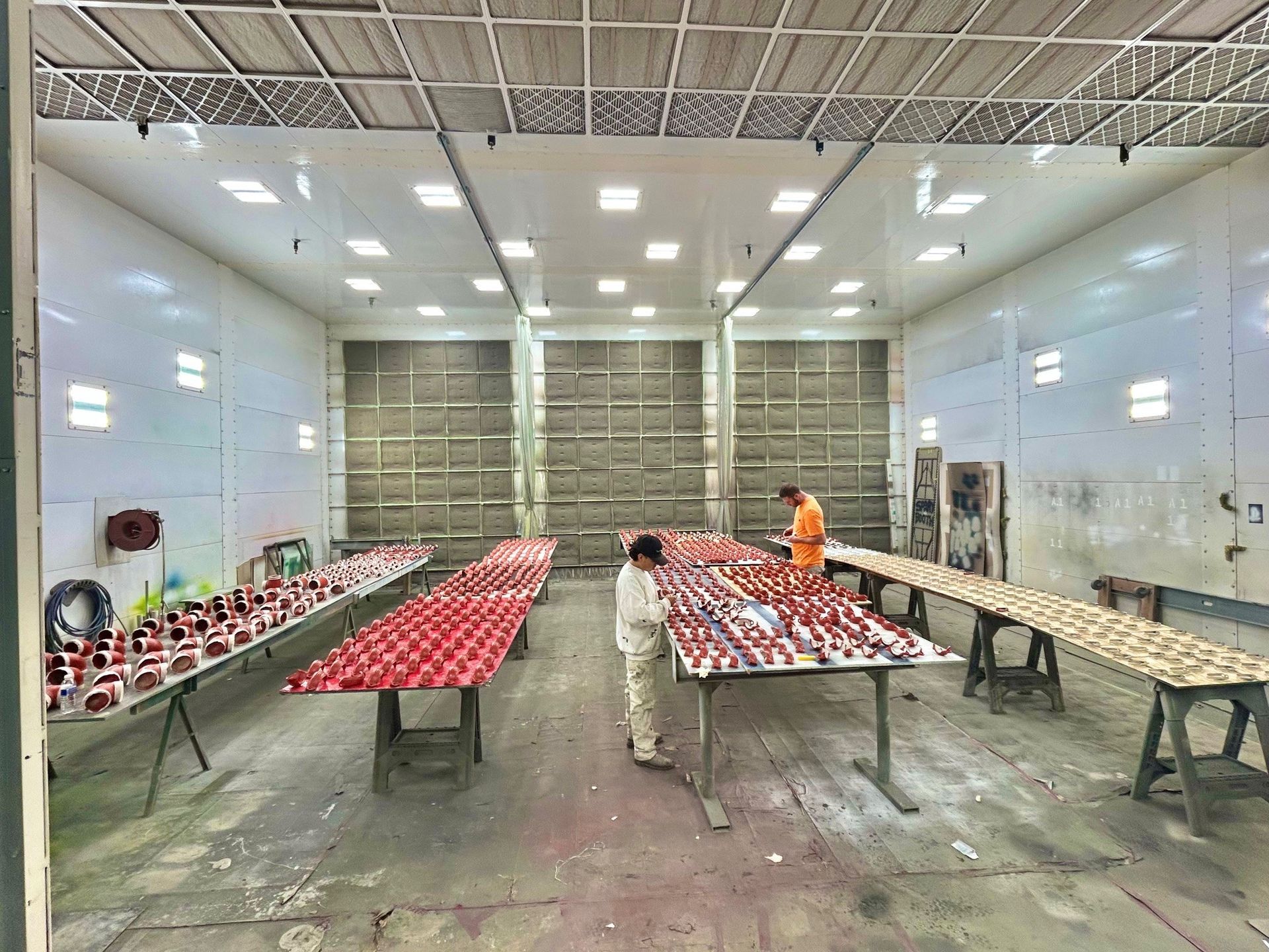 Wide warehouse room with long tables covered in red-and-white items; one worker in orange stands near the back.
