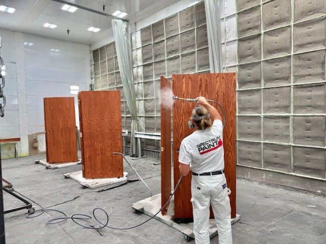 Worker spray-painting wood panels in a workshop booth with ventilation fans and cables on the floor