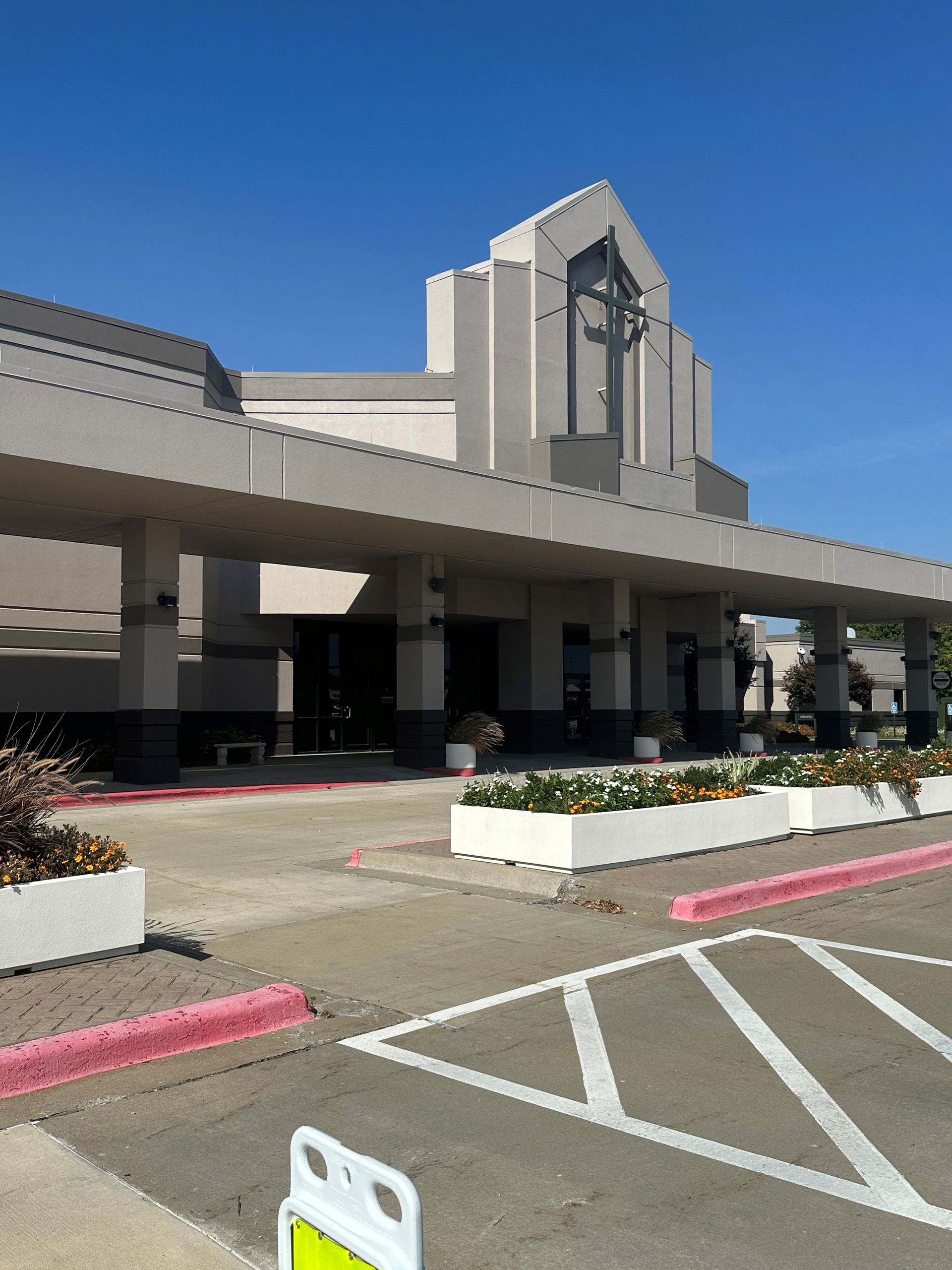 Large beige building entrance with covered drop-off area, white planters, and parking lot under a clear blue sky