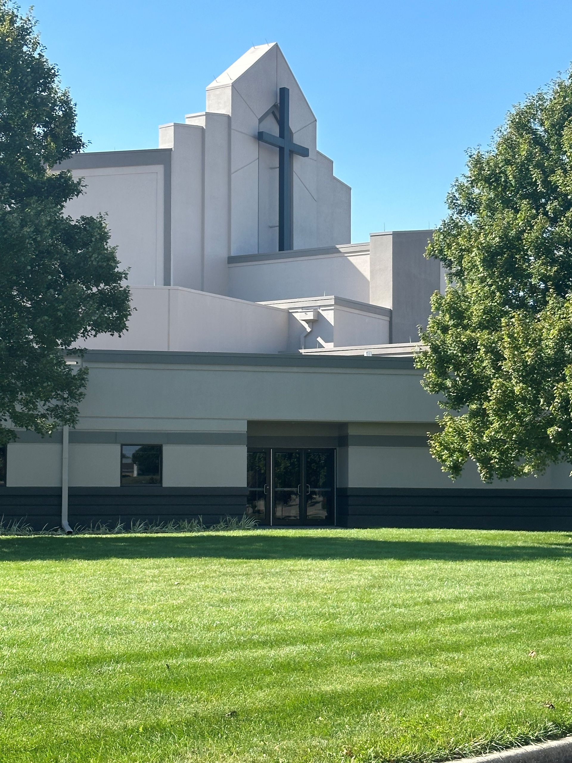 Modern white church building with a cross, framed by trees and a green lawn under a blue sky