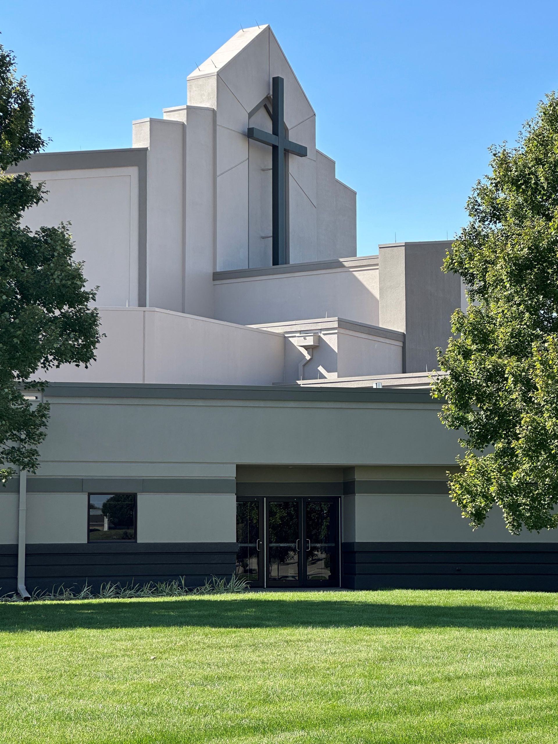 Modern church building with a large cross on the facade, framed by trees and a green lawn.