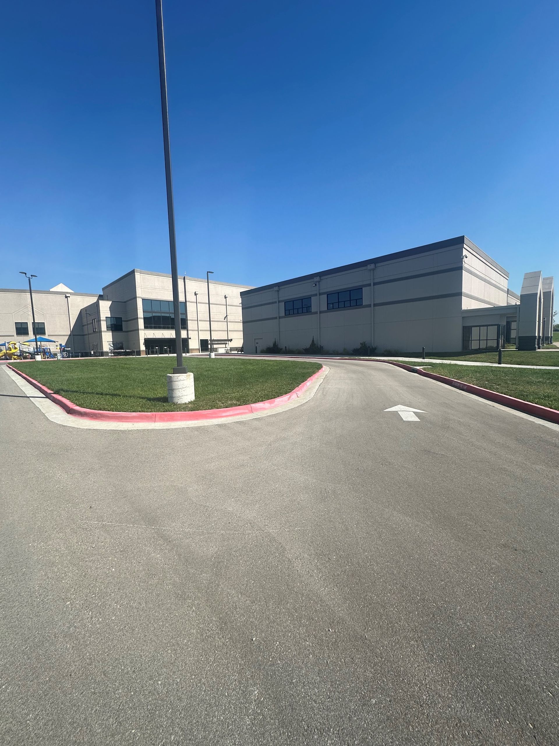 Wide paved road beside beige industrial buildings under a clear blue sky, with a red-curb median and tall pole.