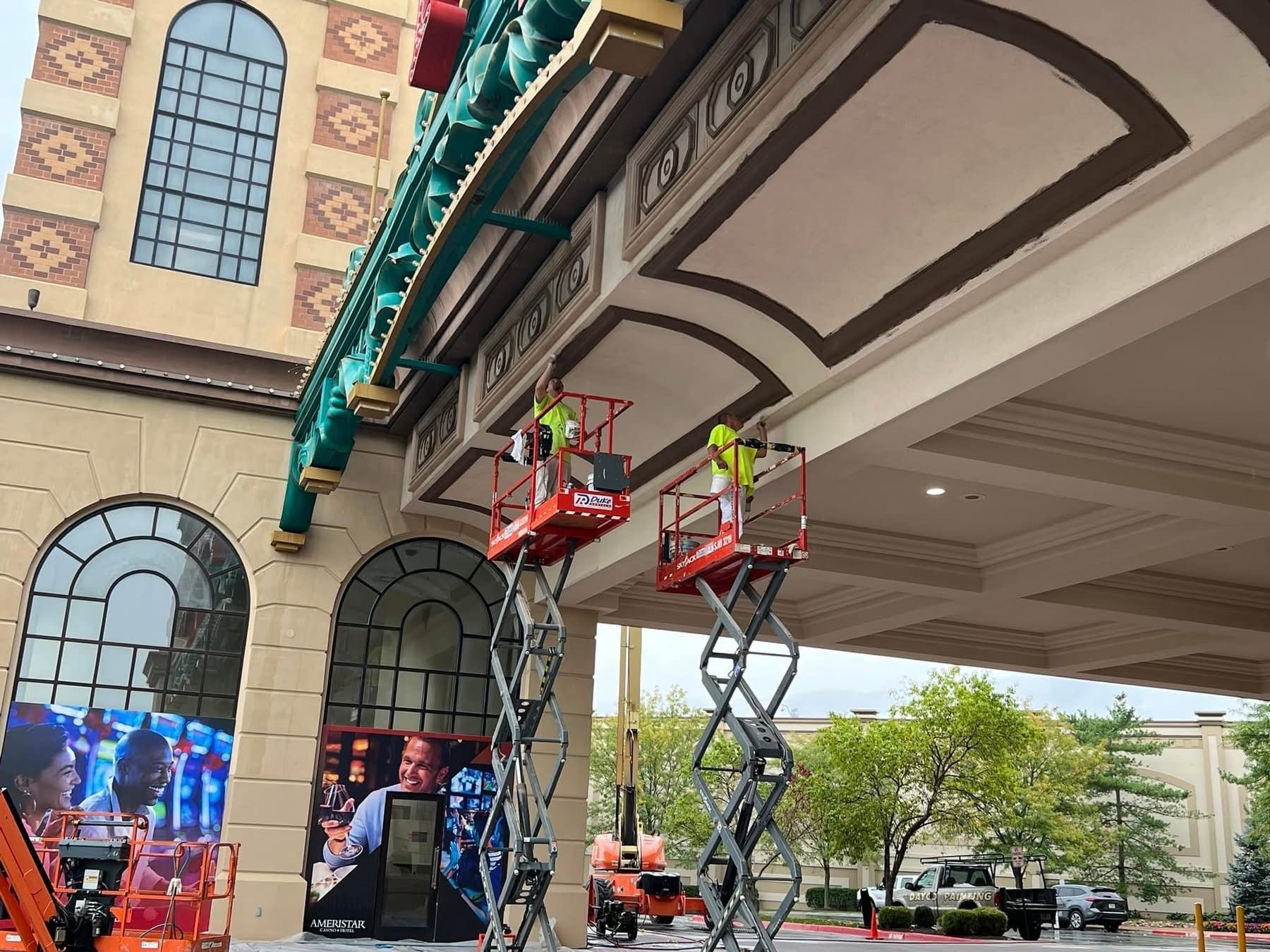 Two workers in scissor lifts under a building awning, inspecting overhead trim and lights.