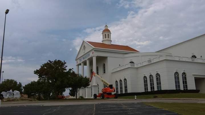 White church building with columns and a red-tiled roof under a cloudy sky, beside a street and parking lot