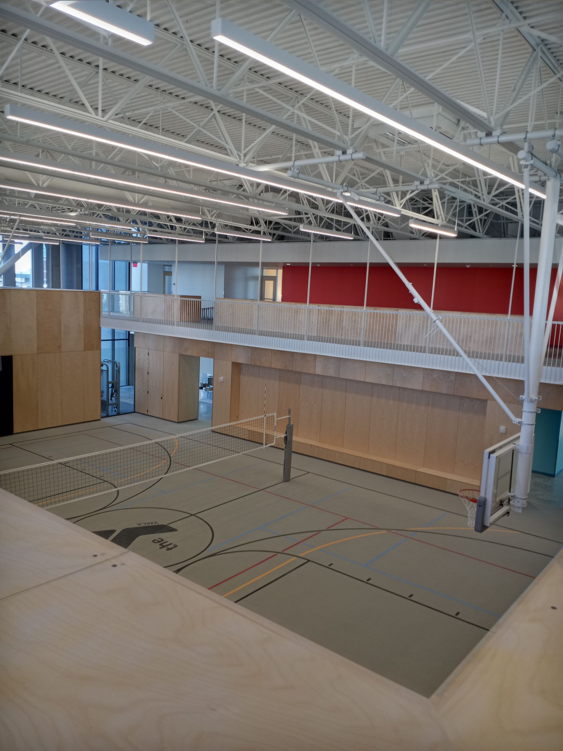 Empty indoor sports court with white ceiling trusses and tan floor mats, red wall panels, and wood-paneled walls