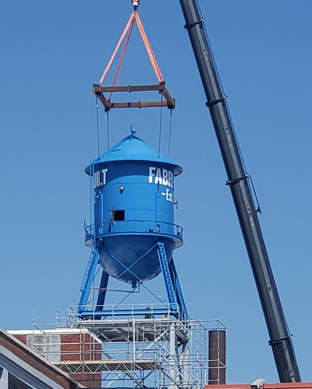Blue water tower being lifted by a crane over a building against a clear sky