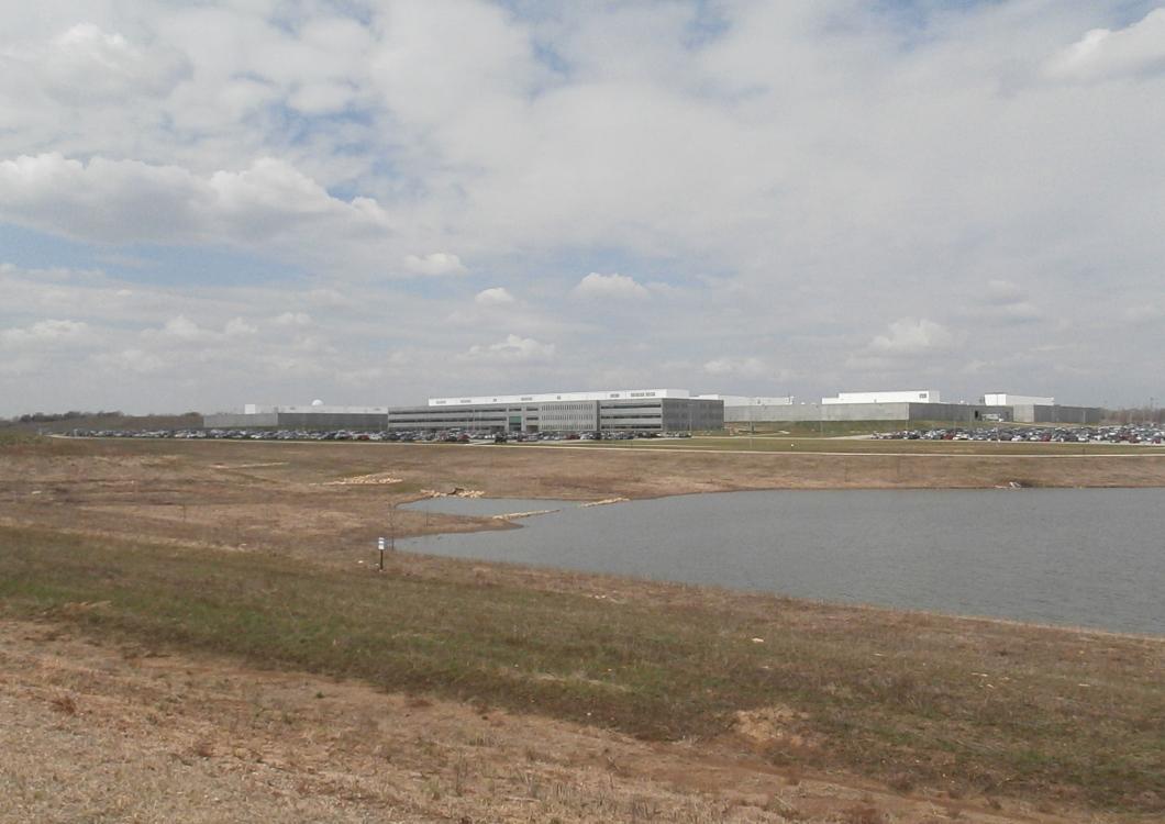 Wide marshland with a small lake under a cloudy sky, with distant buildings on the horizon.