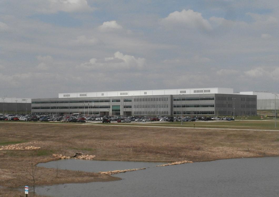 Large gray office building beside a parking lot and grassy field under cloudy skies.