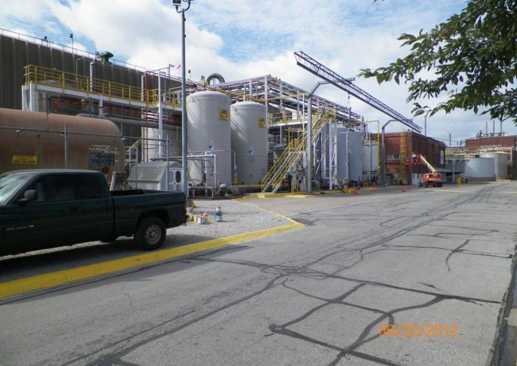 Industrial facility with metal tanks and conveyors beside a cracked road, with a black pickup in the foreground