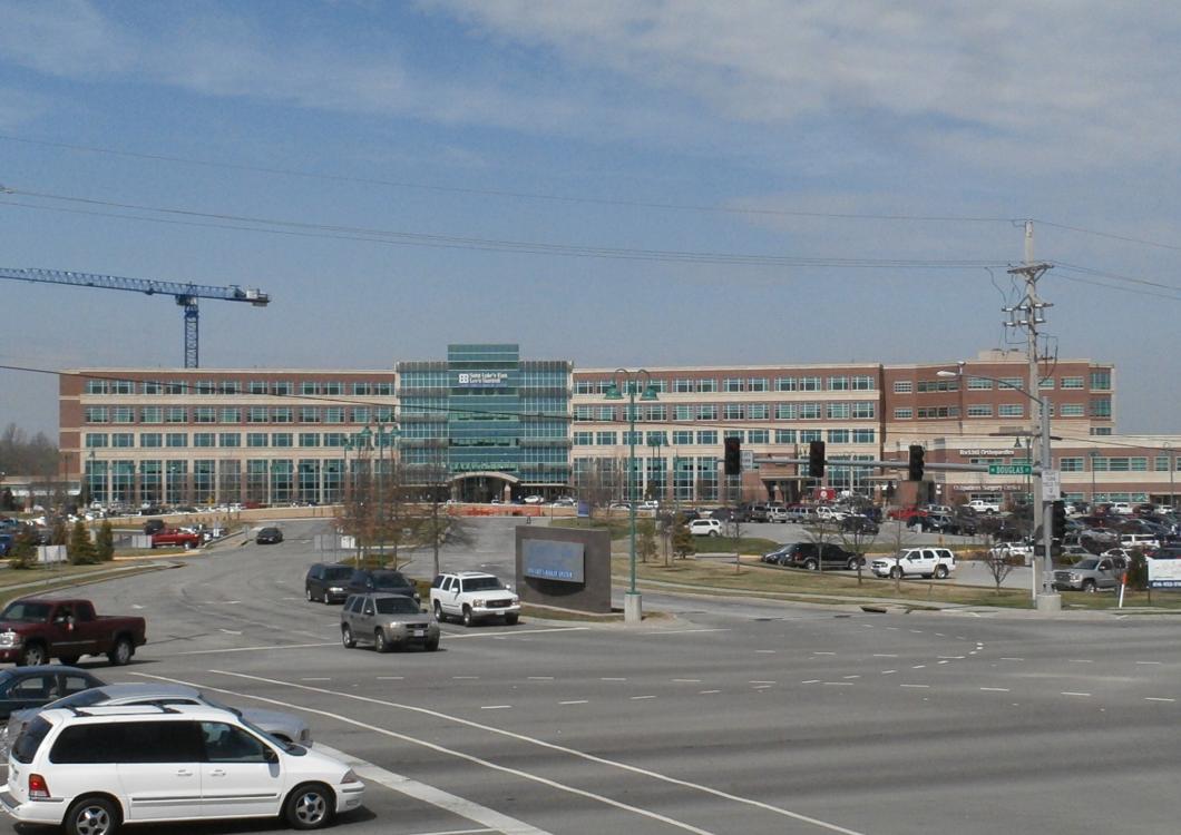 Large modern office building by a busy road, with cars, cranes, and a clear blue sky.
