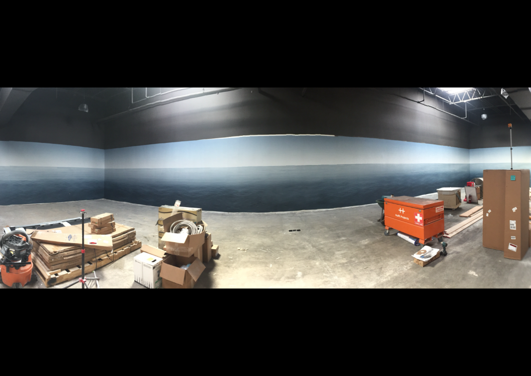 Wide warehouse with a long gray wall, concrete floor, and scattered pallets, boxes, and orange equipment carts.