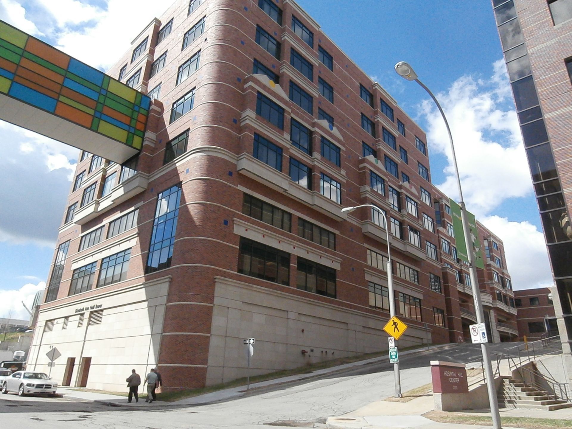 Brick apartment building on a city street corner under a blue sky, with a colorful awning and a few pedestrians.