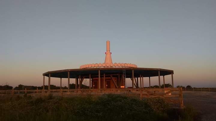 A round pavilion with a central spire at sunset, surrounded by grassy fields and dark support columns.
