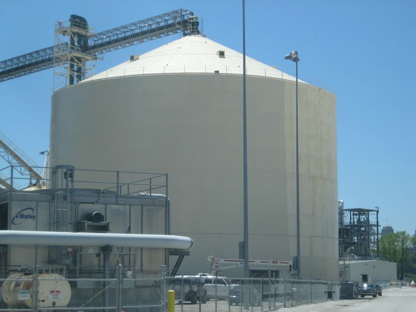 Large white industrial storage tank at a facility, with metal catwalks and piping in the foreground.