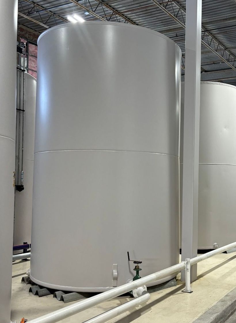 Large white industrial storage tank in a factory with overhead piping and metal ceiling structure