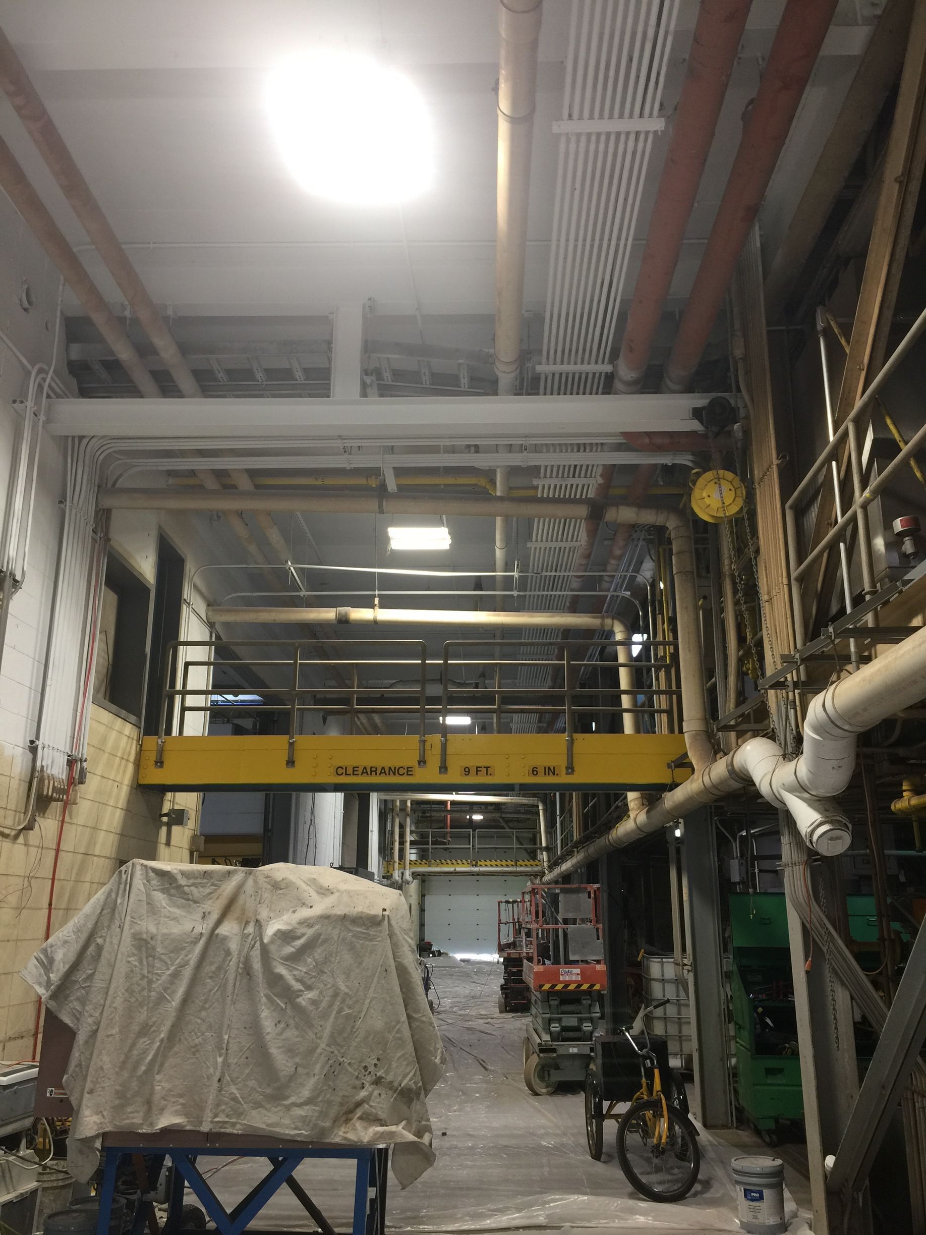 Industrial hallway with exposed pipes, yellow beams, a covered cart, and a bicycle under bright overhead lights
