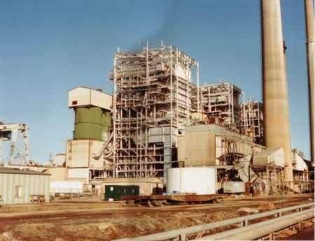 Industrial plant with scaffolding, tall smokestacks, and rail tracks under a clear sky