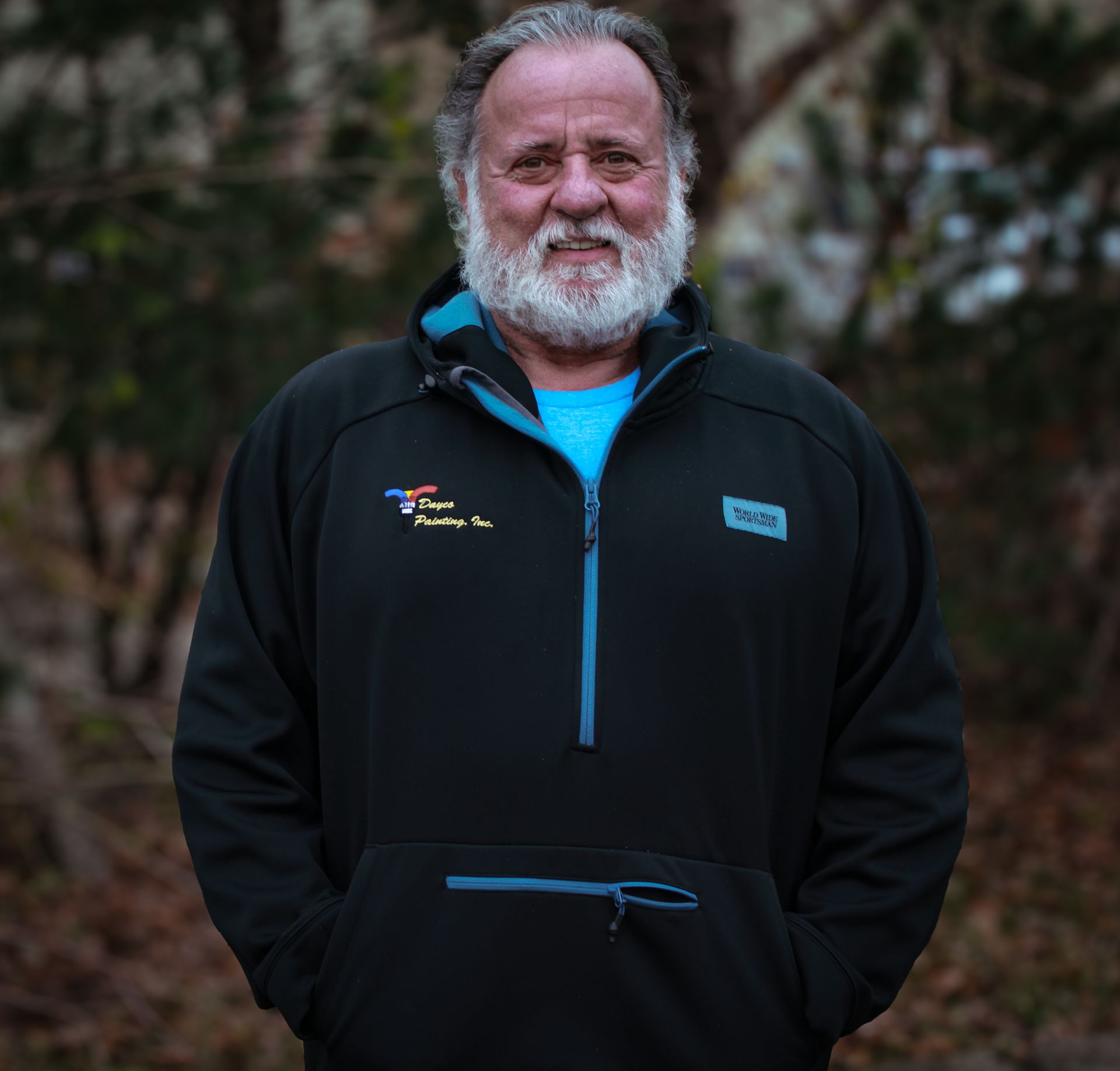 Smiling man in a black jacket with blue trim, standing outdoors against a blurred background