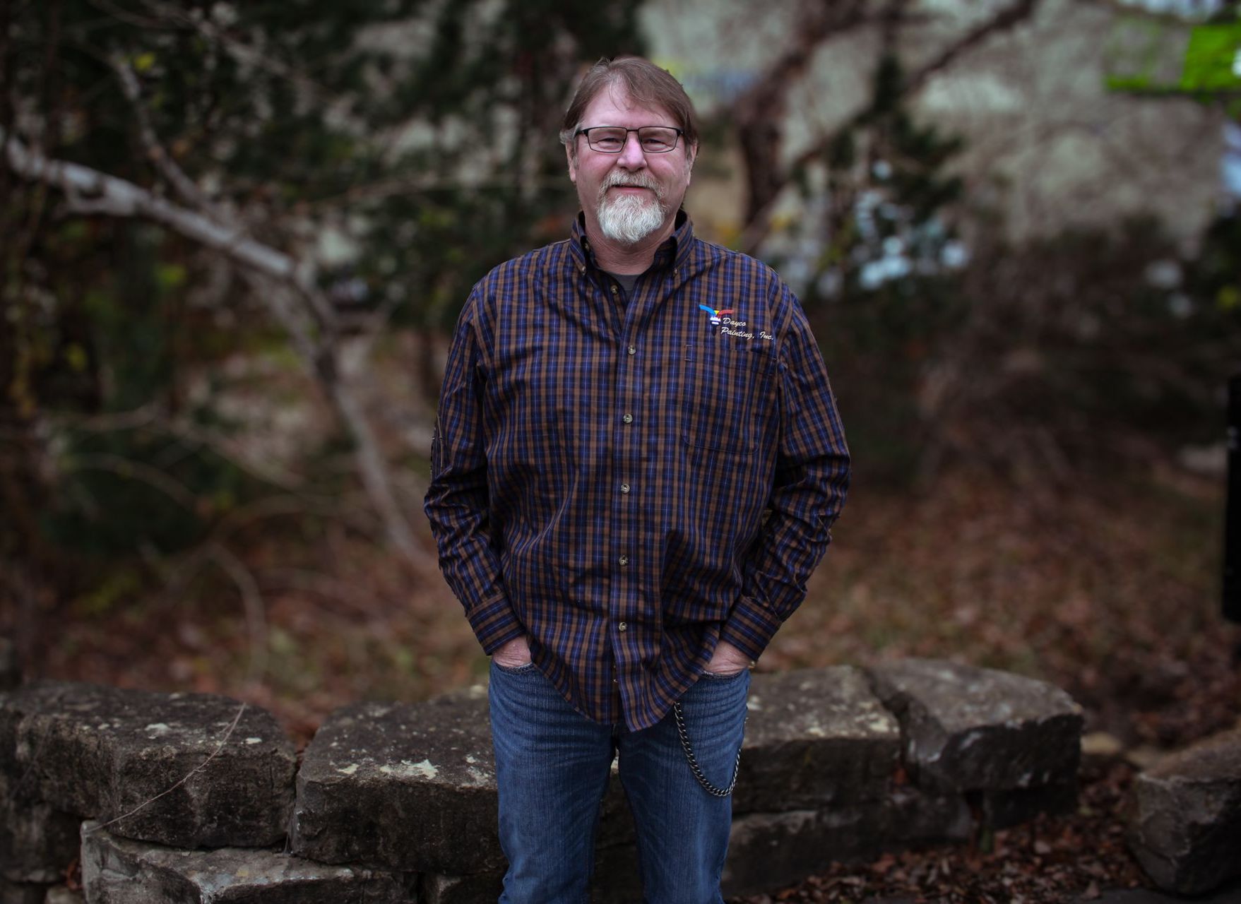 Man standing outdoors in a dark plaid shirt and jeans, in front of rocks and trees.