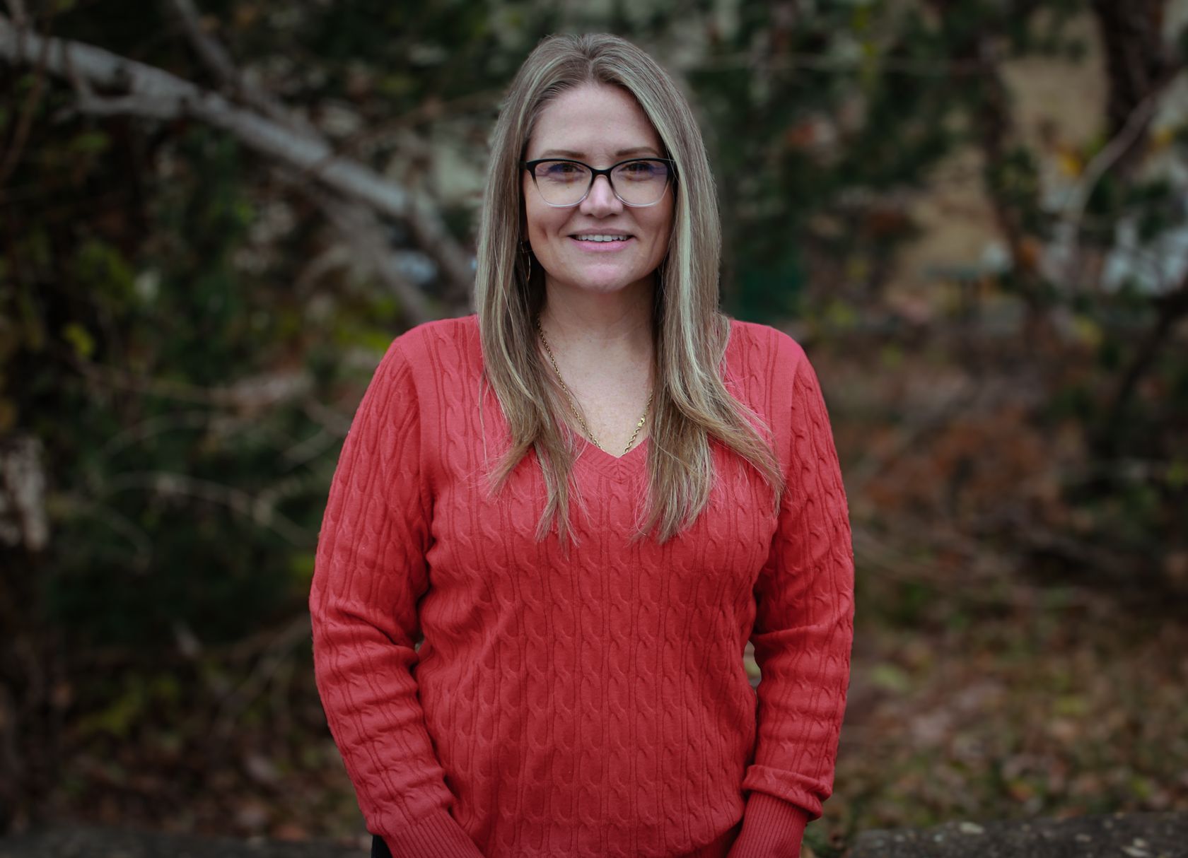 Woman in a red sweater standing outdoors, smiling in front of a blurred wooded background