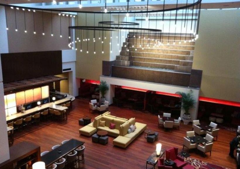 Modern hotel lobby with warm wood floors, central seating area, and hanging pendant lights under a high ceiling