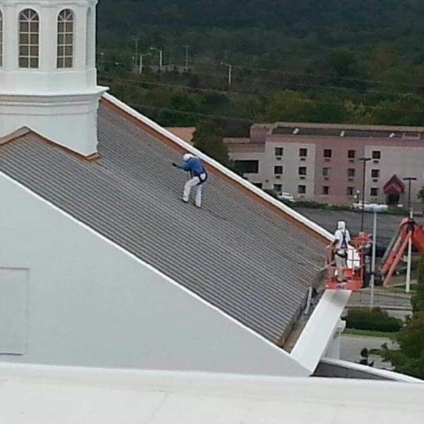 Worker in blue jacket on a steep church roof, with equipment and another worker below.