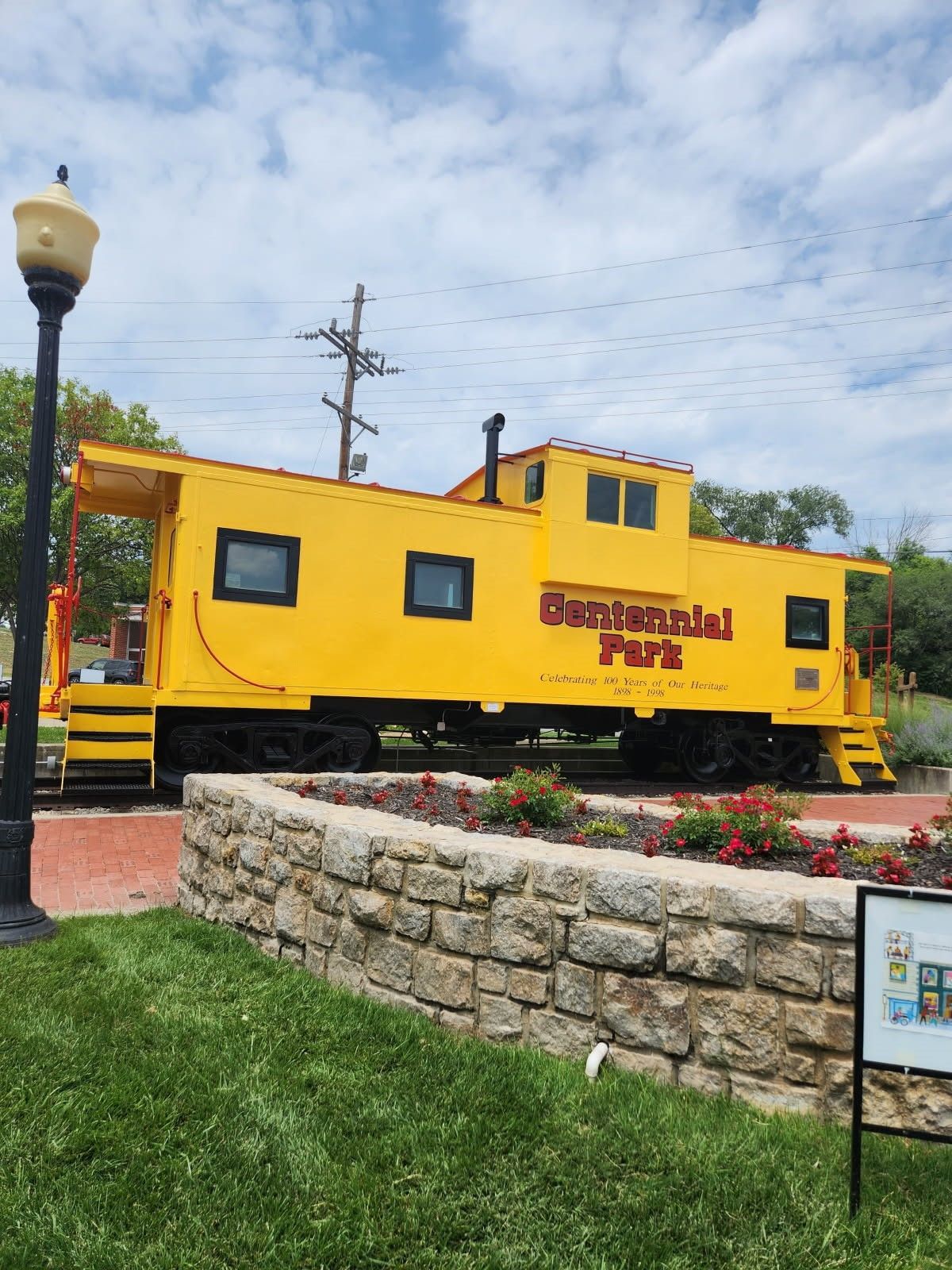 Yellow caboose-style train car on display beside a stone wall and flowers in a park.