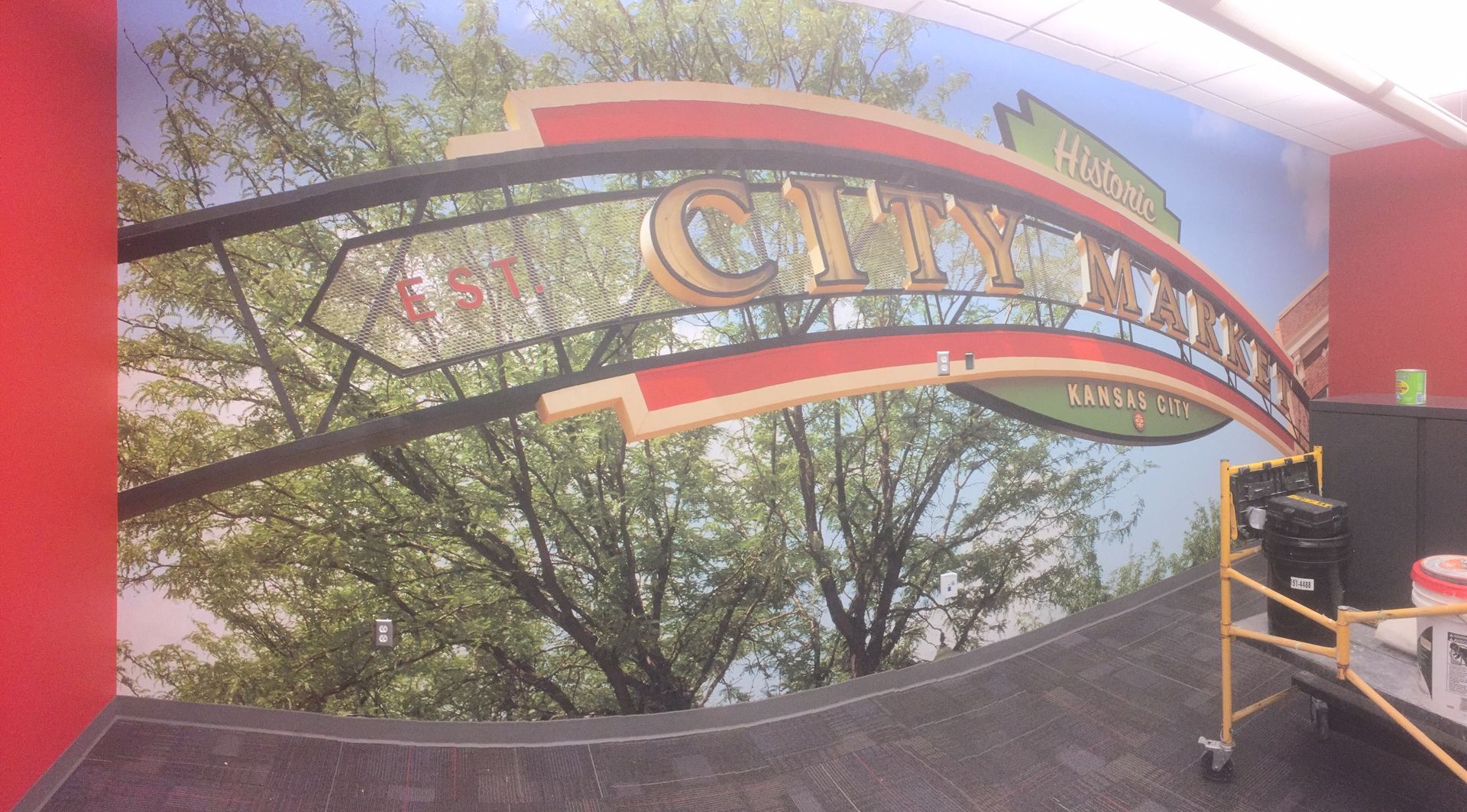 Colorful arched sign over a walkway with trees in the background and a red wall on the left