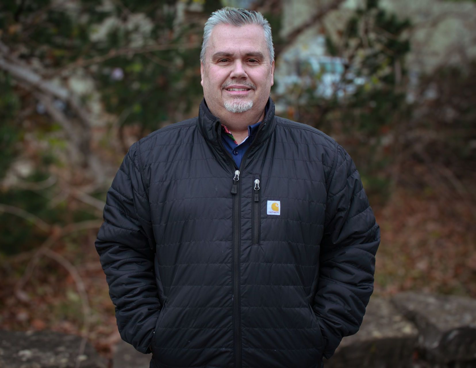 Man in a black jacket standing outdoors with hands in pockets, looking at the camera.