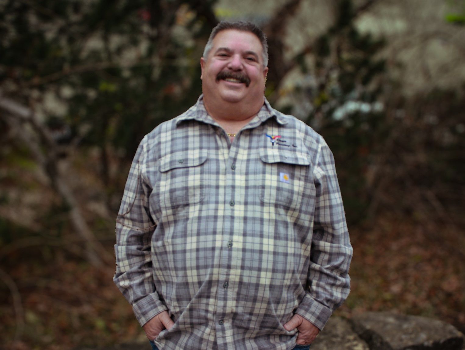 Man in a plaid shirt smiling outdoors with hands in pockets, standing in front of a blurred forest background.