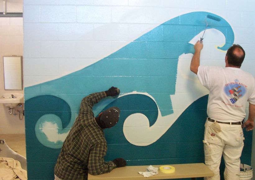 Two people painting a blue wave mural on a tiled wall.
