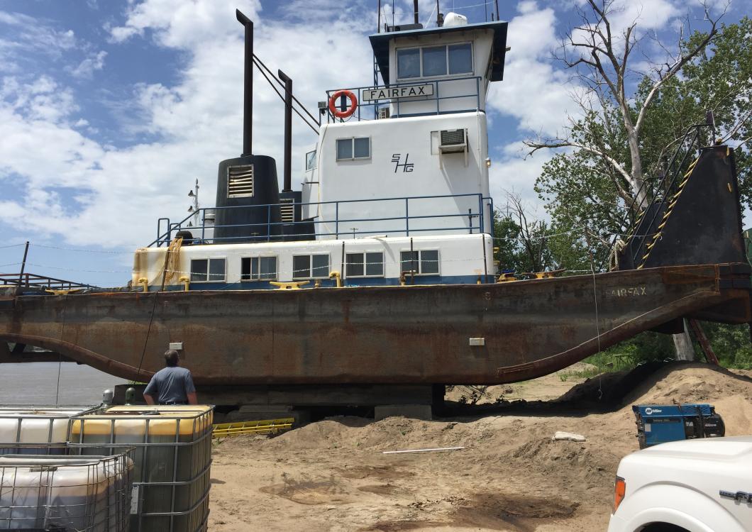 Rusty tugboat on a sandy shore, with a person nearby and a white truck in the foreground.