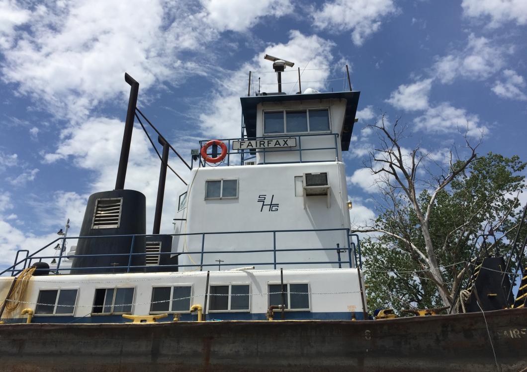 White tugboat with a black smokestack under a blue sky with clouds, seen from the side.