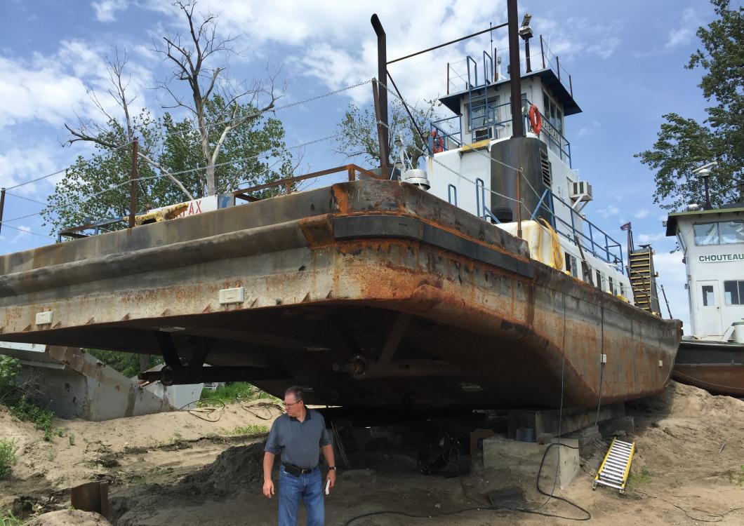 Large rusted boat on land with a man standing underneath it near the shore