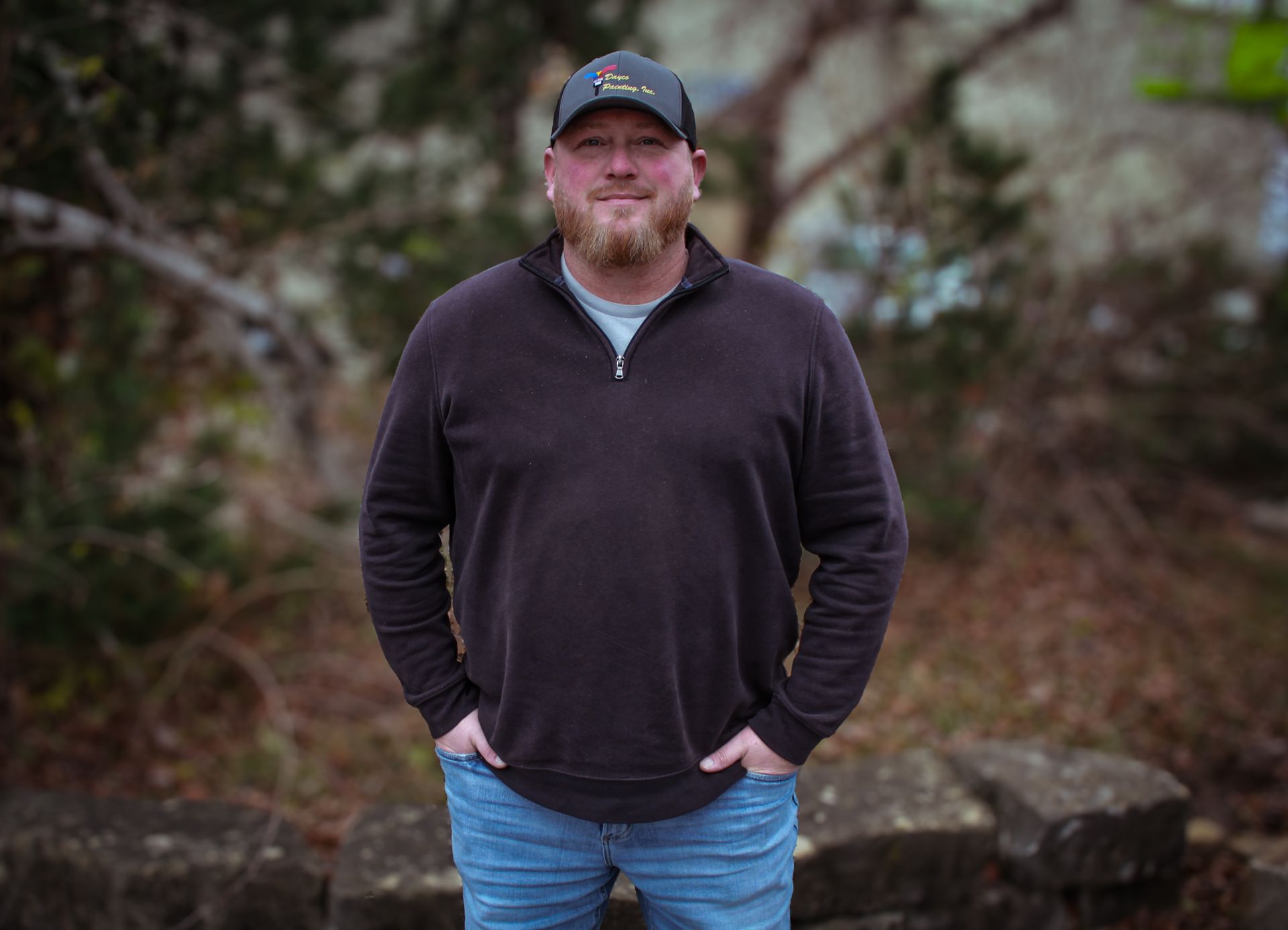 Man standing outdoors in a dark pullover and jeans, hands in pockets, with blurred foliage behind him