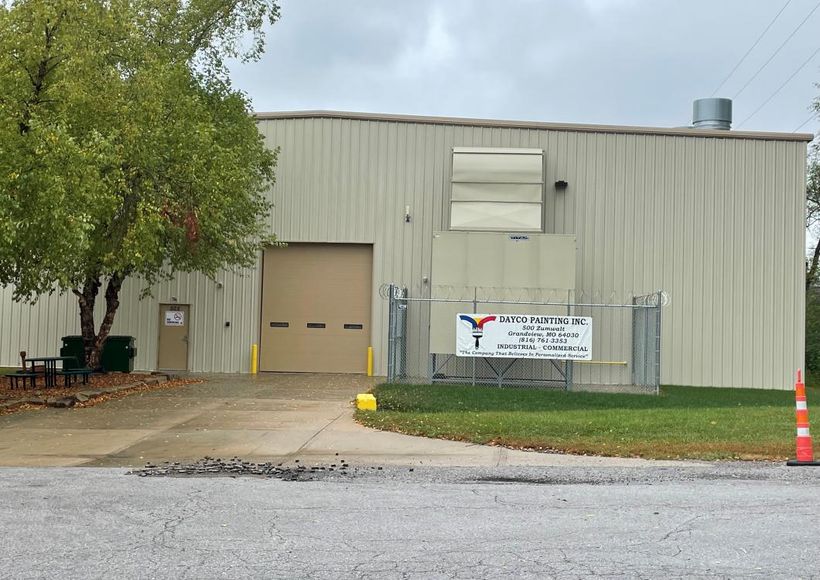 Industrial building with beige exterior, metal fence, tree, and gravel driveway under cloudy sky