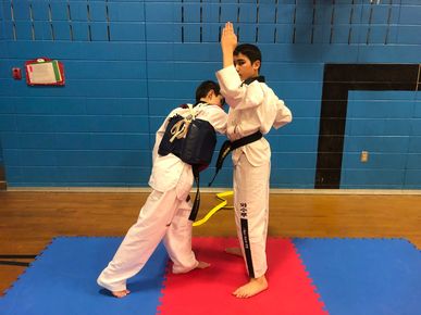 Deux pratiquants d'arts martiaux s'entraînent au combat sur des tapis bleus, l'un frappant vers le haut tandis que l'autre bloque, dans une salle de sport avec un mur bleu.