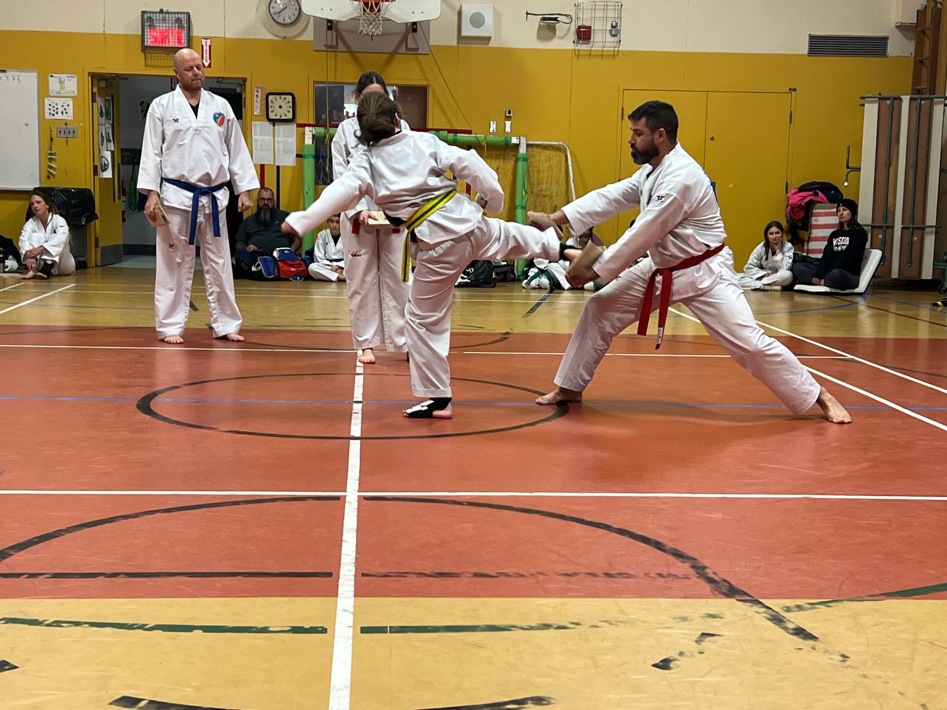 Deux pratiquants d'arts martiaux s'entraînent au combat sur des tapis bleus et rouges dans une salle de sport, levant les mains pour bloquer ou frapper.