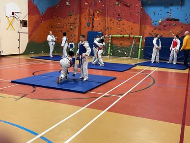 Des enfants pratiquent l'escrime dans un gymnase sur des tapis bleus, avec un mur d'escalade et un panier de basket en arrière-plan.