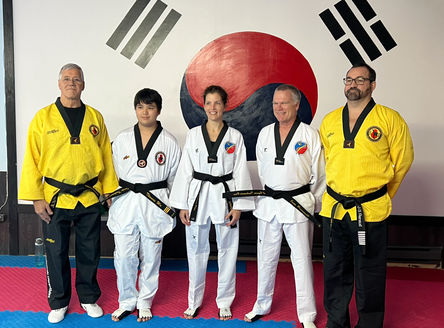 Cinq pratiquants d’arts martiaux en tenue blanche et jaune posent dans un studio de taekwondo devant un drapeau coréen