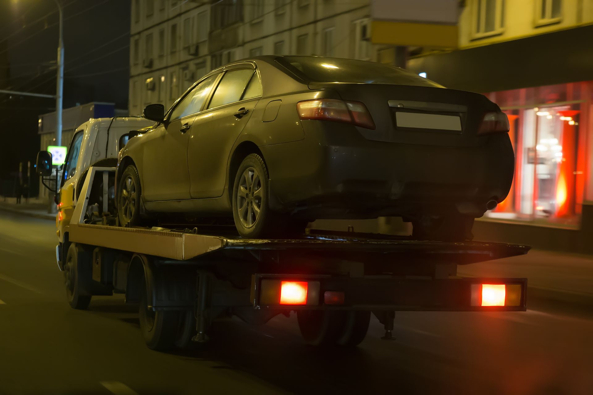 Flatbed tow truck carrying a gray sedan along a dimly lit city street at night.