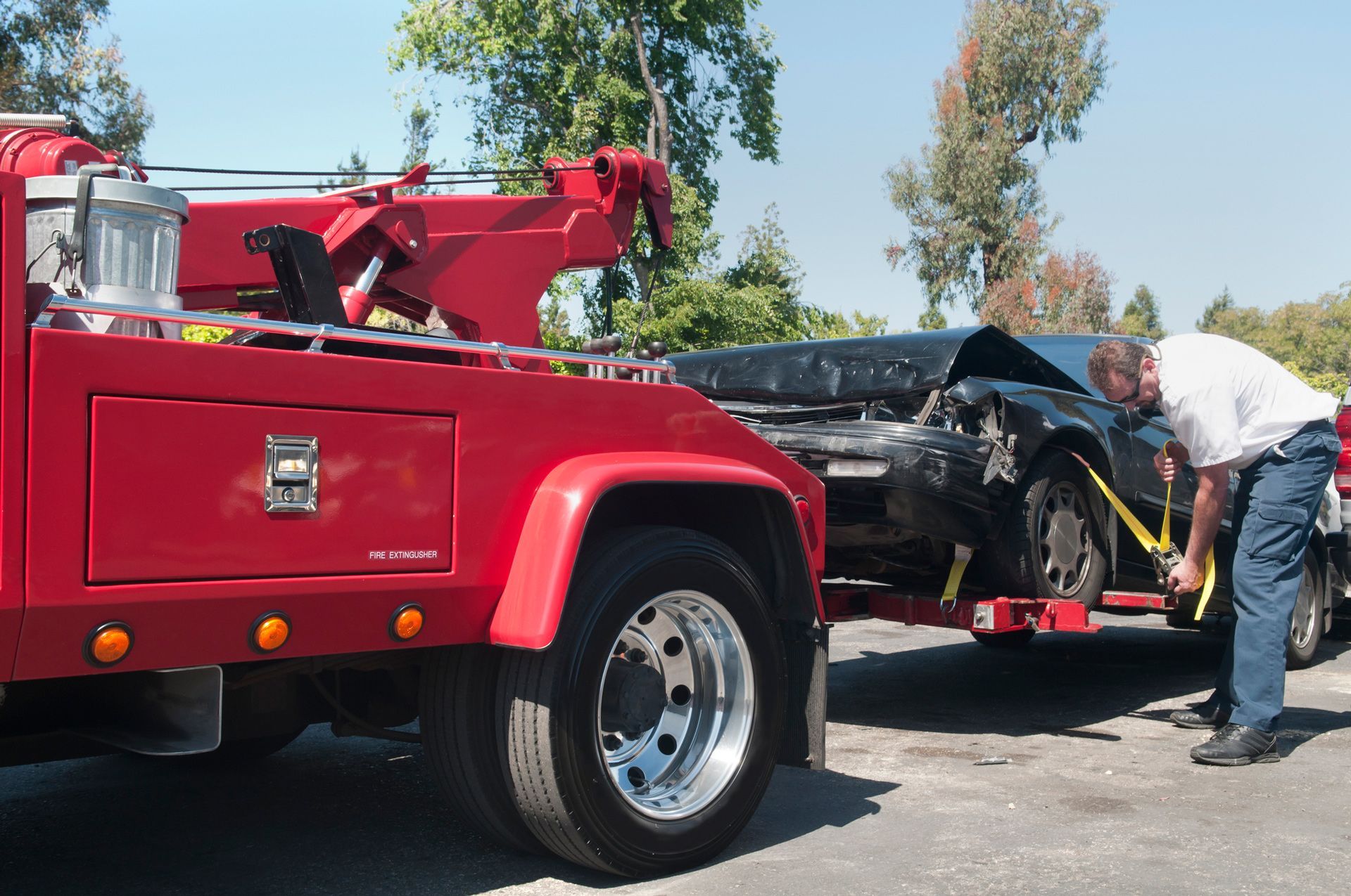 Red tow truck loading a heavily damaged black car on a sunny day. Man securing the vehicle.