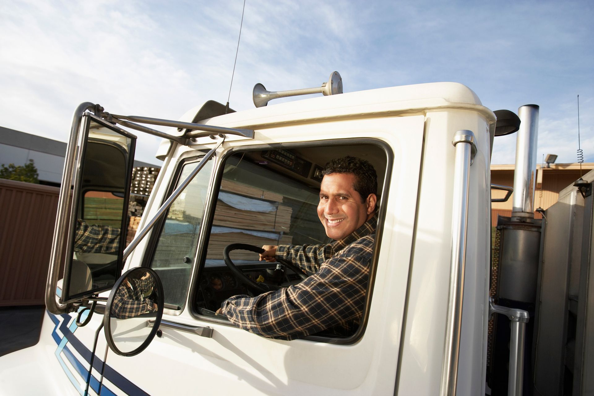 Smiling man in a plaid shirt driving a white semi-truck on a sunny day.