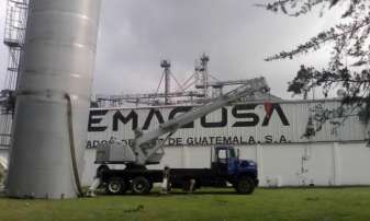 Un tractor azul y un camión blanco estacionados junto a un gran tanque industrial y un edificio con el letrero “EMACUSA”.