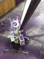 Un pequeño robot con brazos morados y ruedas blancas está sentado sobre un suelo de hormigón bajo una estructura metálica.
