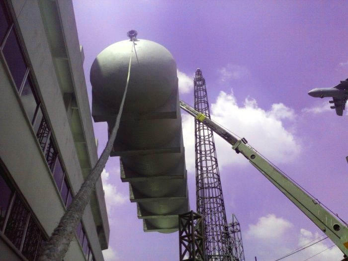 Un gran tanque cilíndrico plateado es izado por una grúa junto a un edificio bajo un cielo púrpura.