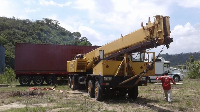 Un camión grúa amarillo levanta un contenedor de transporte rojo en un terreno baldío, con un trabajador cerca y colinas al fondo.